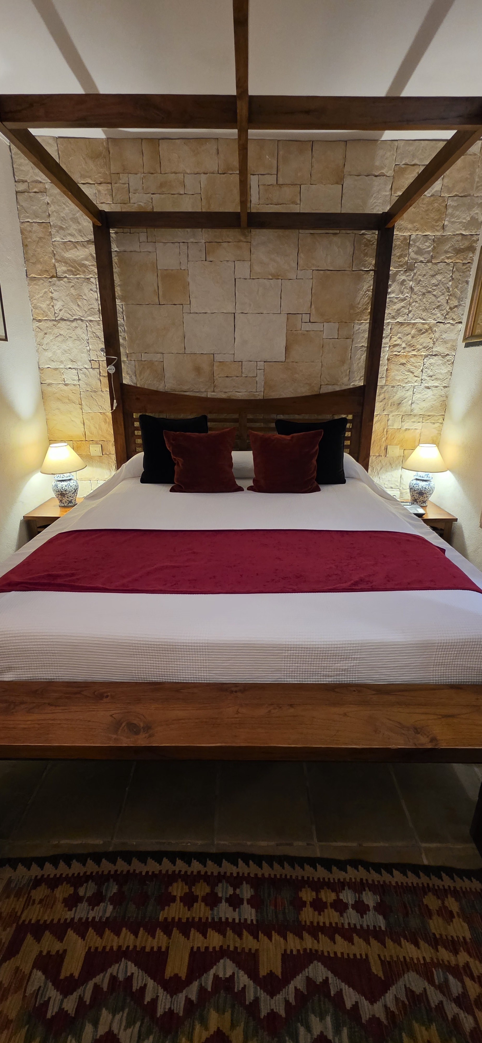 A four-poster bed with deep-red cover against a bare stone wall, kilim at its foot