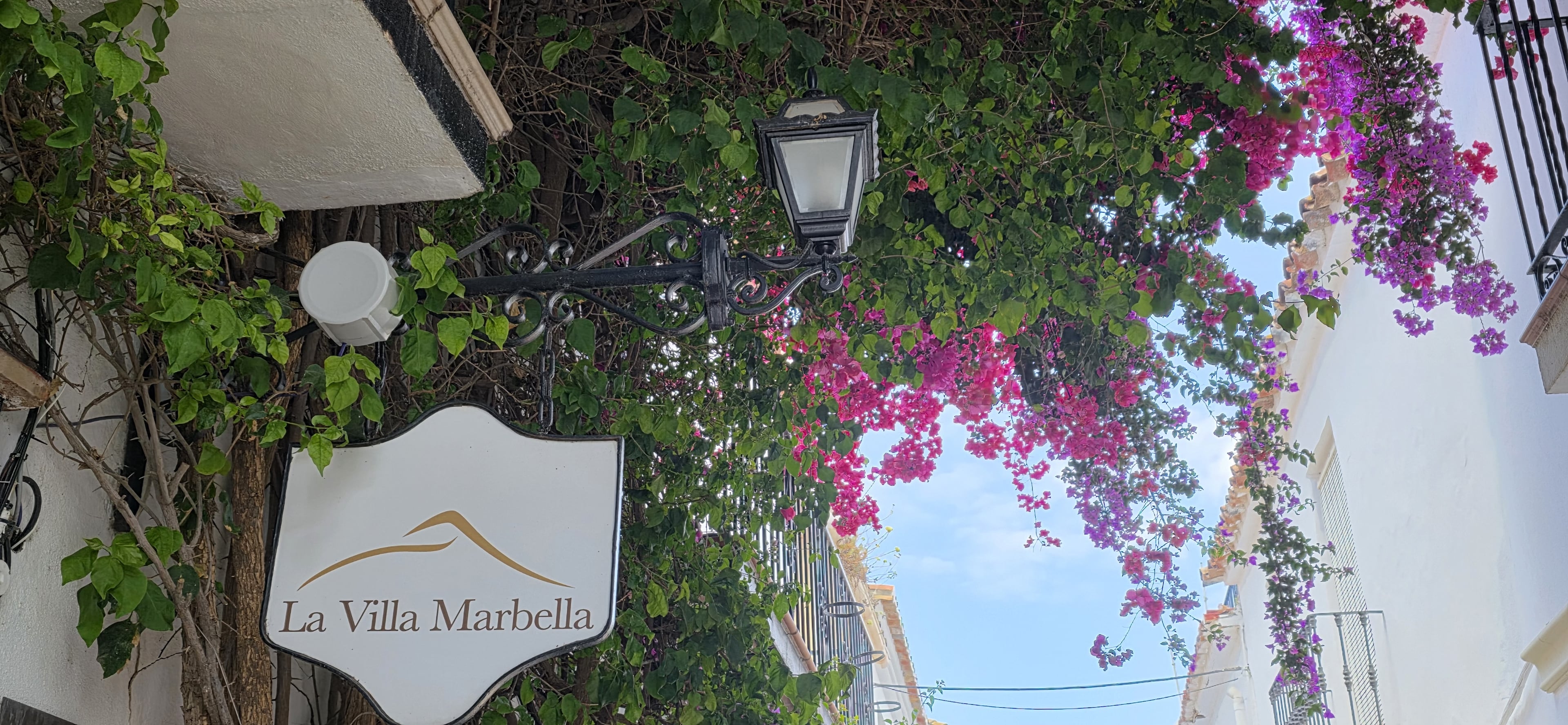 The hanging La Villa Marbella sign and wrought-iron lantern on a whitewashed old-town lane, framed by bougainvillea against the sky