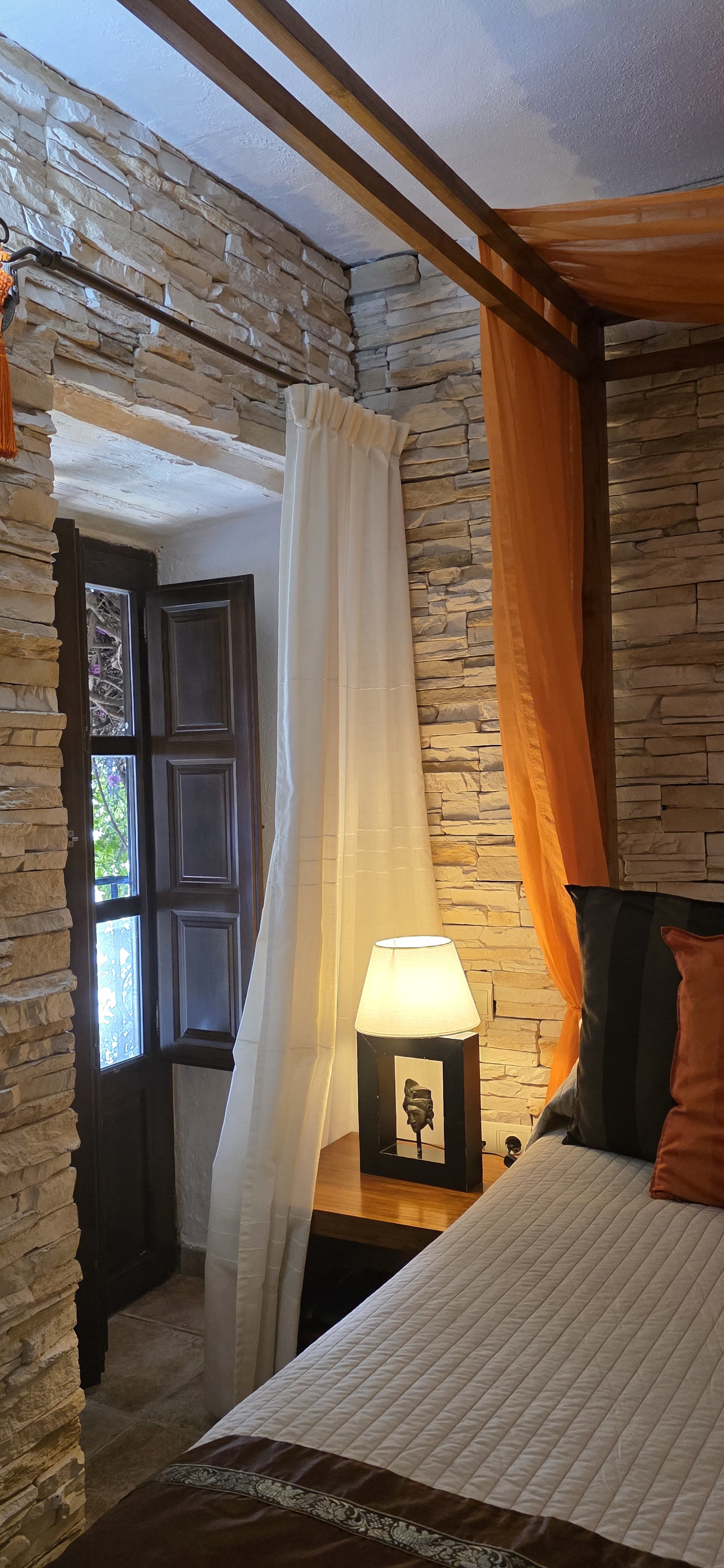 Four-poster bed with saffron canopy beside a shuttered window and stone wall